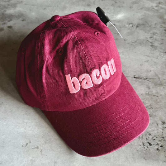 A burgundy cotton baseball cap with the word 'bacon' embroidered in light pink across the front, resting on a gray concrete surface.