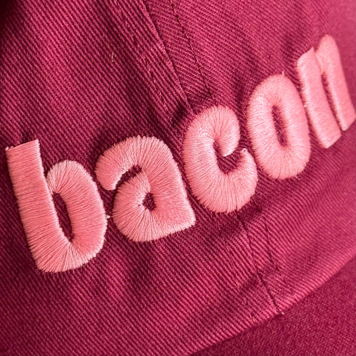 A close-up of a dark pink or magenta fabric surface with light pink embroidered letters spelling 'bacon' 