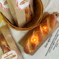 Baguette candle lit on a wood board. Baguette candles in a bag in a basket next to it.
