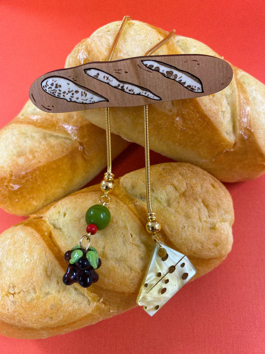Bolo tie with a horizontal baguette as the main piece and smaller charms of olives, grapes and cheese below 