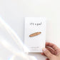 Enamel pin of a baguette with a smiley face, on top of a white card backing that says "it's a pin!" Hand holding the card.