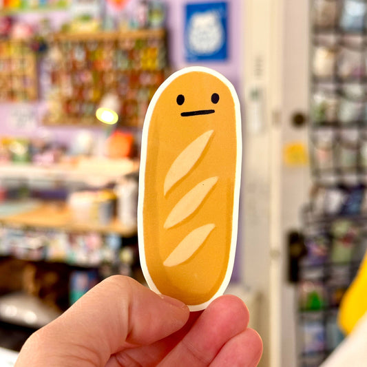 Hand holding a cartoon-style bread-shaped sticker with a store in the background