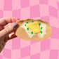 A hand holds a hair claw clip shaped like a potato, featuring a beige skin, white creamy interior, a yellow center, and green speckles, set against a pink checkered background with the brand name 'SARAH PILAR' at the bottom.