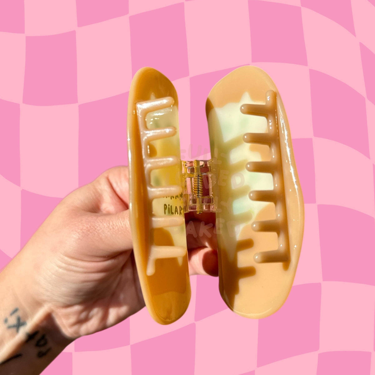 A hand holds a large, glossy baked potato-shaped hair claw clip against a pink and light pink checkerboard background. The brand name 'SARAH PILAR' is printed at the bottom of the image.
