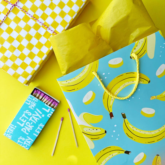 staged table top featuring a banana gift bag with yellow tissue. Checkered yellow tissue and matches on the table as well.