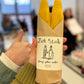 Set of two yellow beeswax taper candles shaped like fish. Shown in paper wrapping with "Fish Sticks" branding and logo. Package is held in a person's hand for scale.
