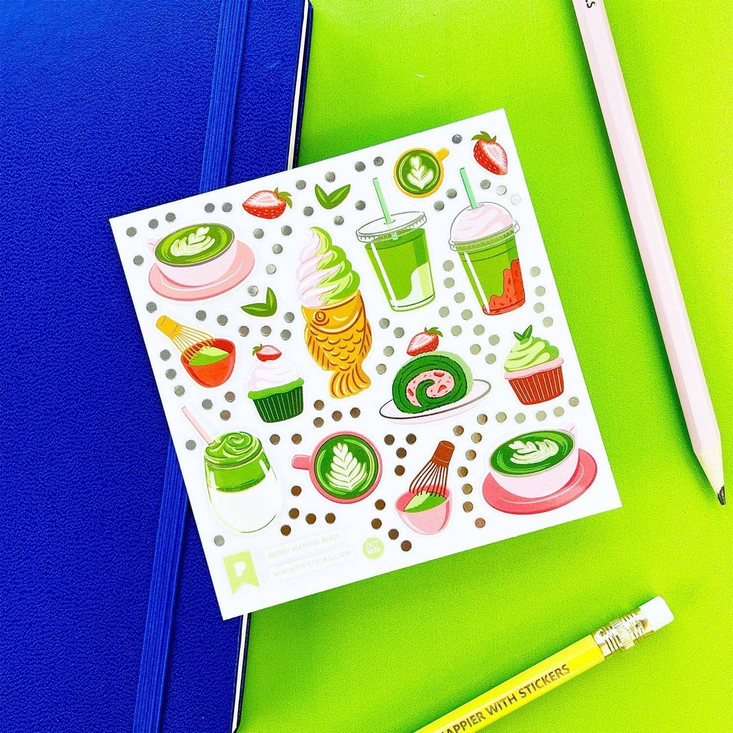 Sheet of colorful matcha food-themed stickers on a green surface with a blue notebook and white pen.