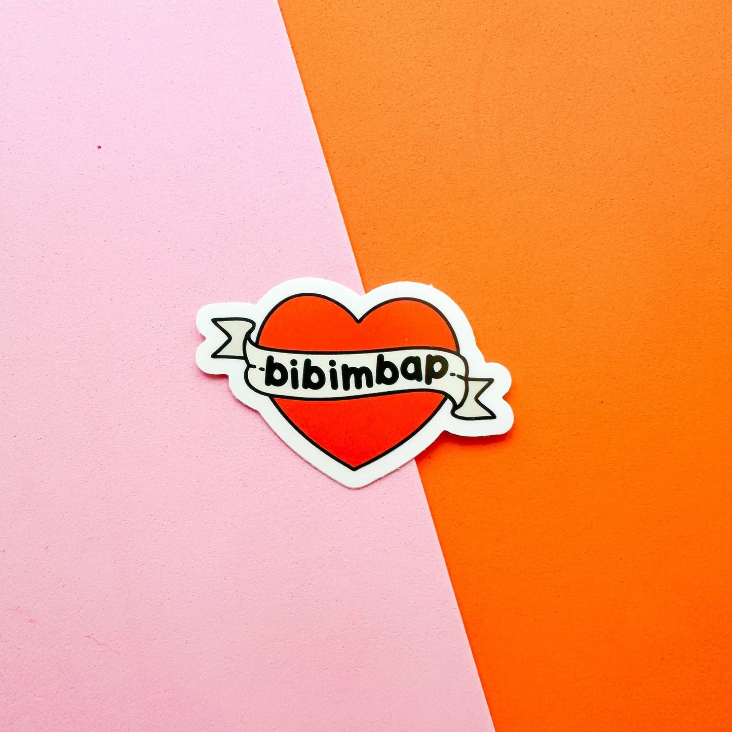 A die-cut sticker featuring a red heart with a white border and a banner across it that reads 'bibimbap' in black text, placed on a diagonally split pink and orange background.