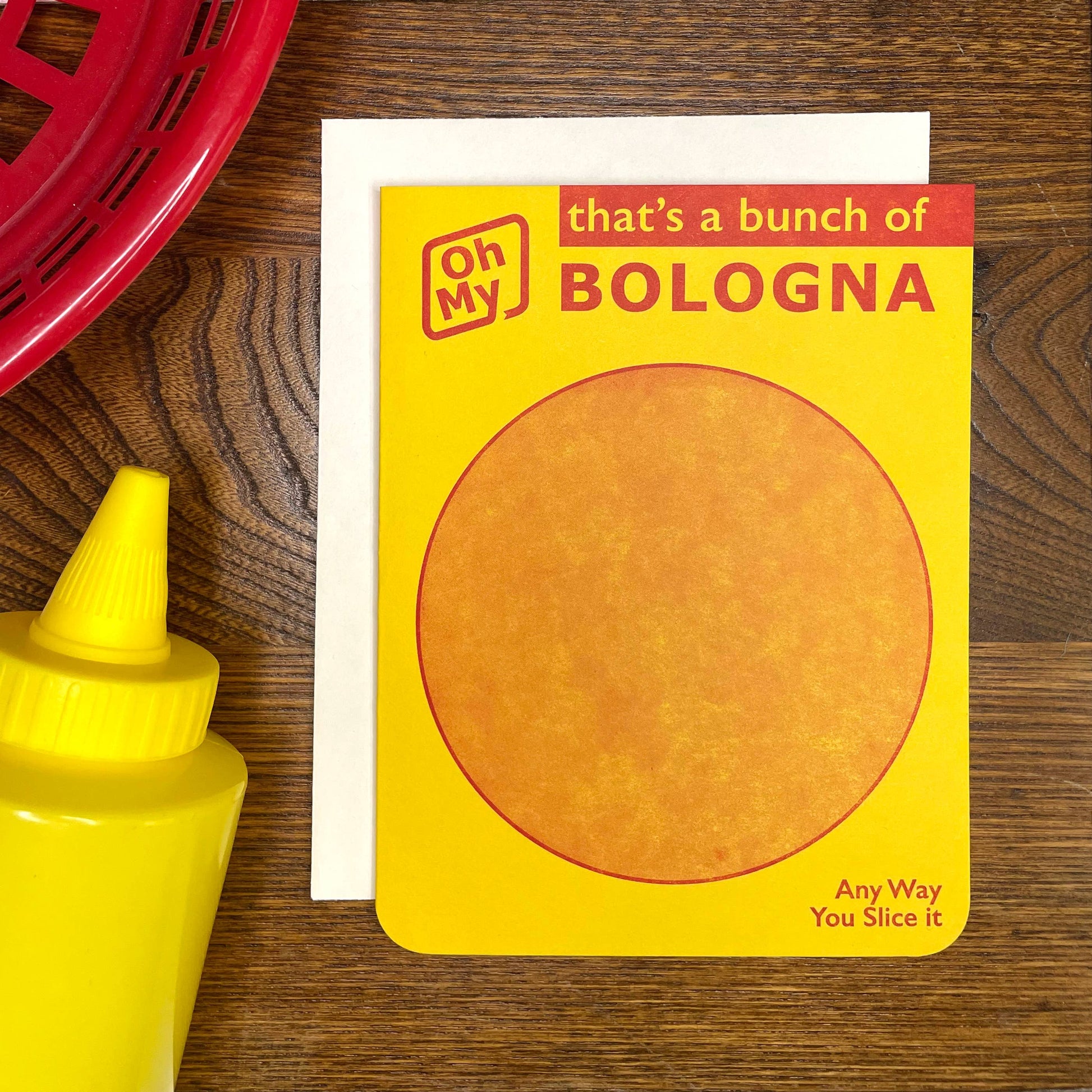 Greeting card with a bologna illustration and text on a wooden surface with a mustard bottle and red basket.