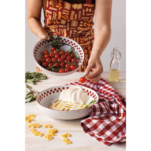 A person wearing a patterned apron holds a bowl of cherry tomatoes above a white bowl with sliced mozzarella and pasta on a light wood table, with a red-and-white checkered cloth and a glass bottle nearby.
