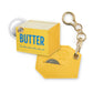 A yellow faux-leather keychain wallet shaped like a butter carton and a matching hexagonal tag, both attached to gold-tone hardware and a white key ring, on a white background.