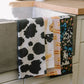 Butter tea towel shown with other printed towels hanging over the side of a kitchen sink.