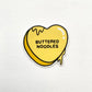 Heart-shaped yellow sticker with 'BUTTERED NOODLES' text on a white background