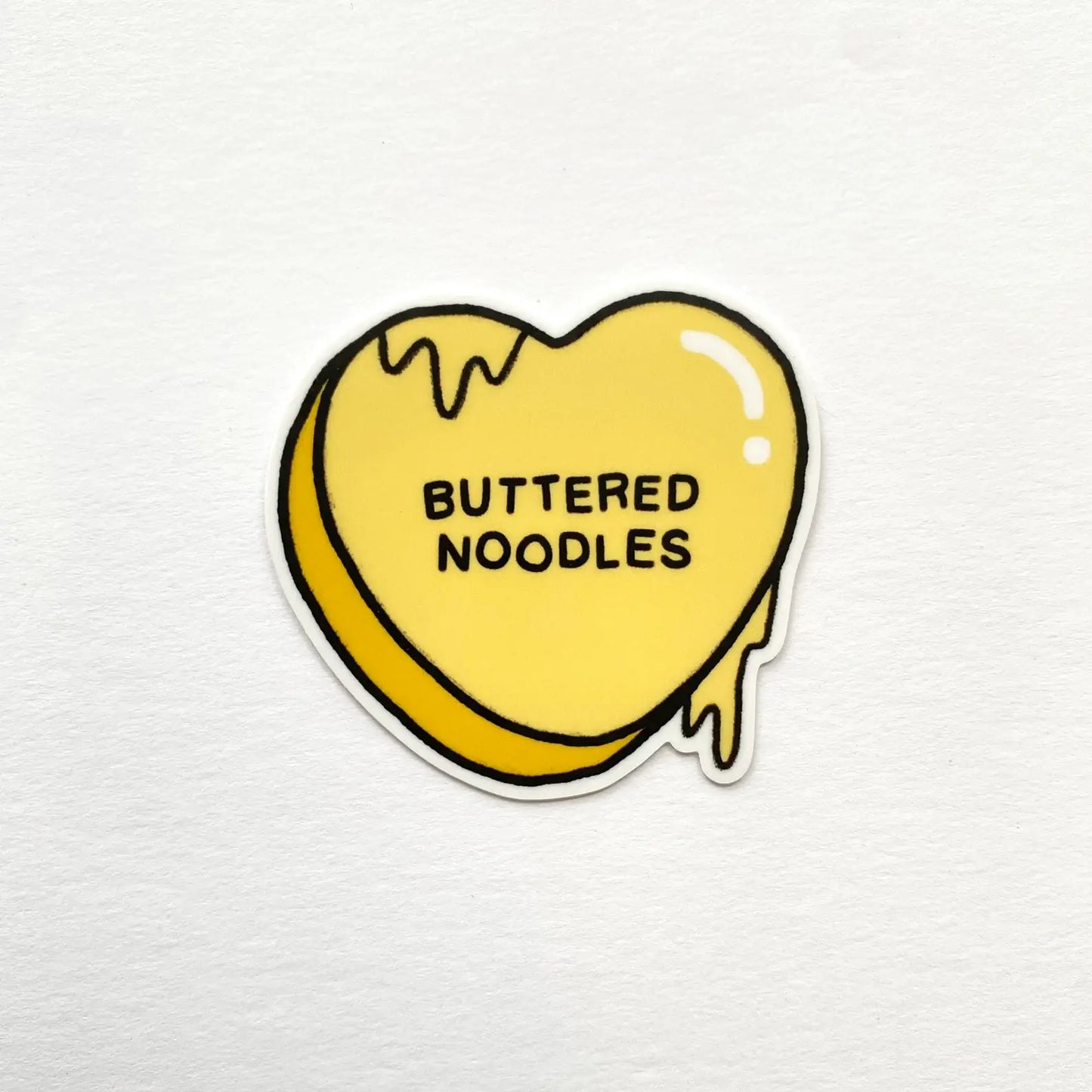 Heart-shaped yellow sticker with 'BUTTERED NOODLES' text on a white background