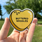 Heart-shaped sticker with 'Buttered Noodles' text held in a hand outdoors