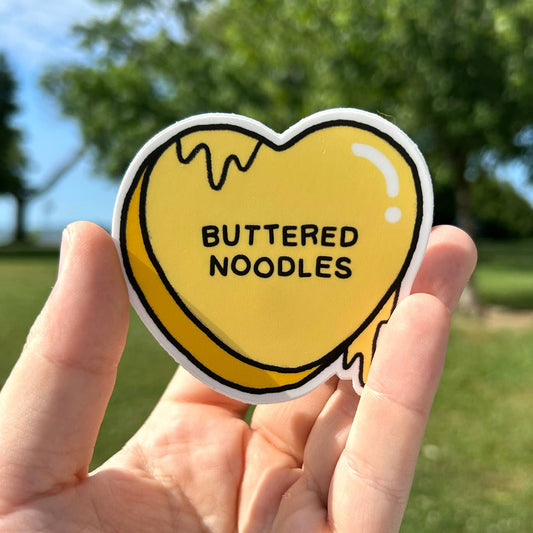 Heart-shaped sticker with 'Buttered Noodles' text held in a hand outdoors