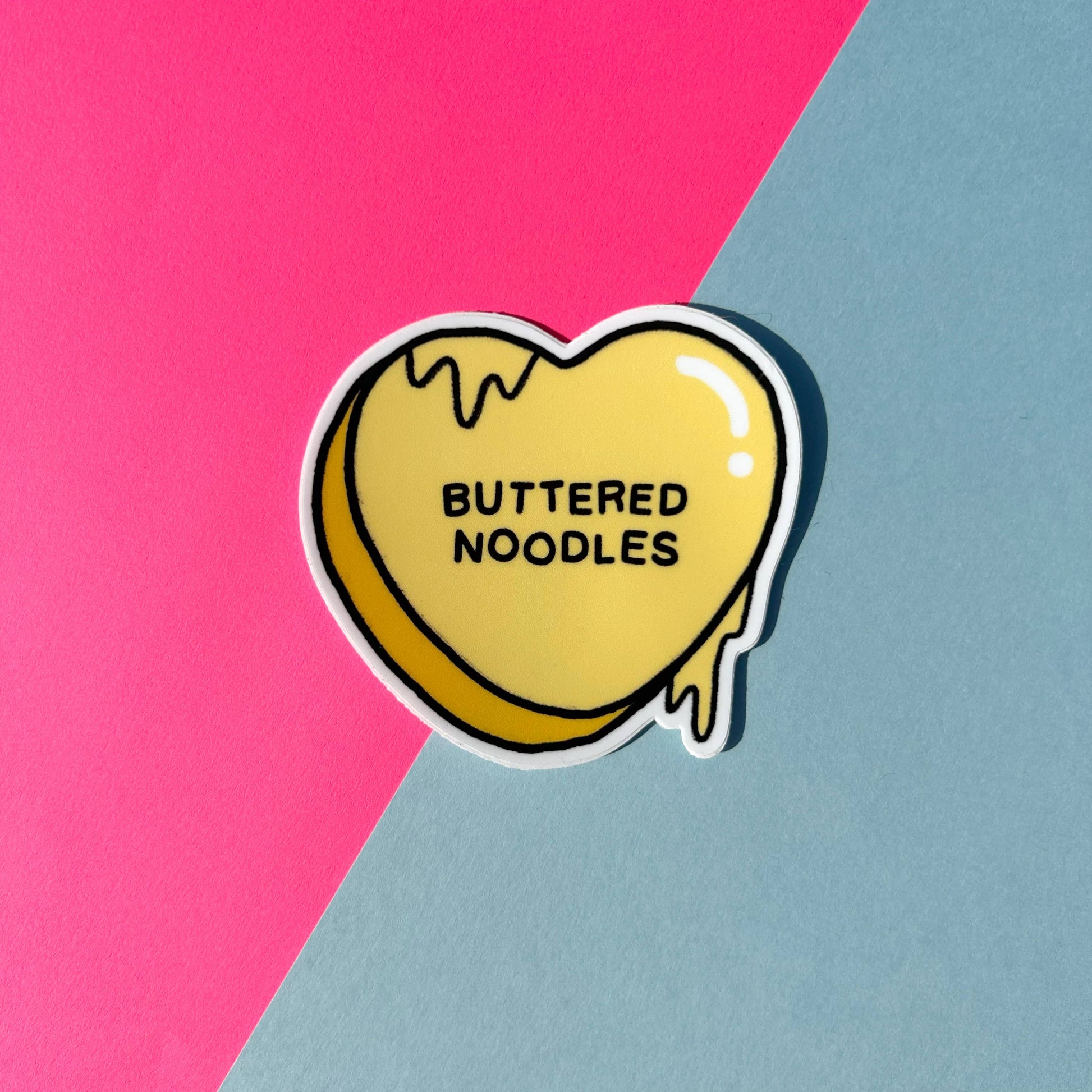 Heart-shaped 'Buttered Noodles' candy sticker on a pink and blue background