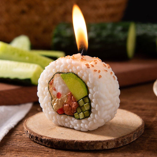 A novelty sushi roll candle shaped like a maki roll, with white rice, green avocado, and crab filling details, topped with sesame seeds and a small flame burning from the top. It sits on a round wooden slice coaster on a wooden table with cucumber slices blurred in the background.