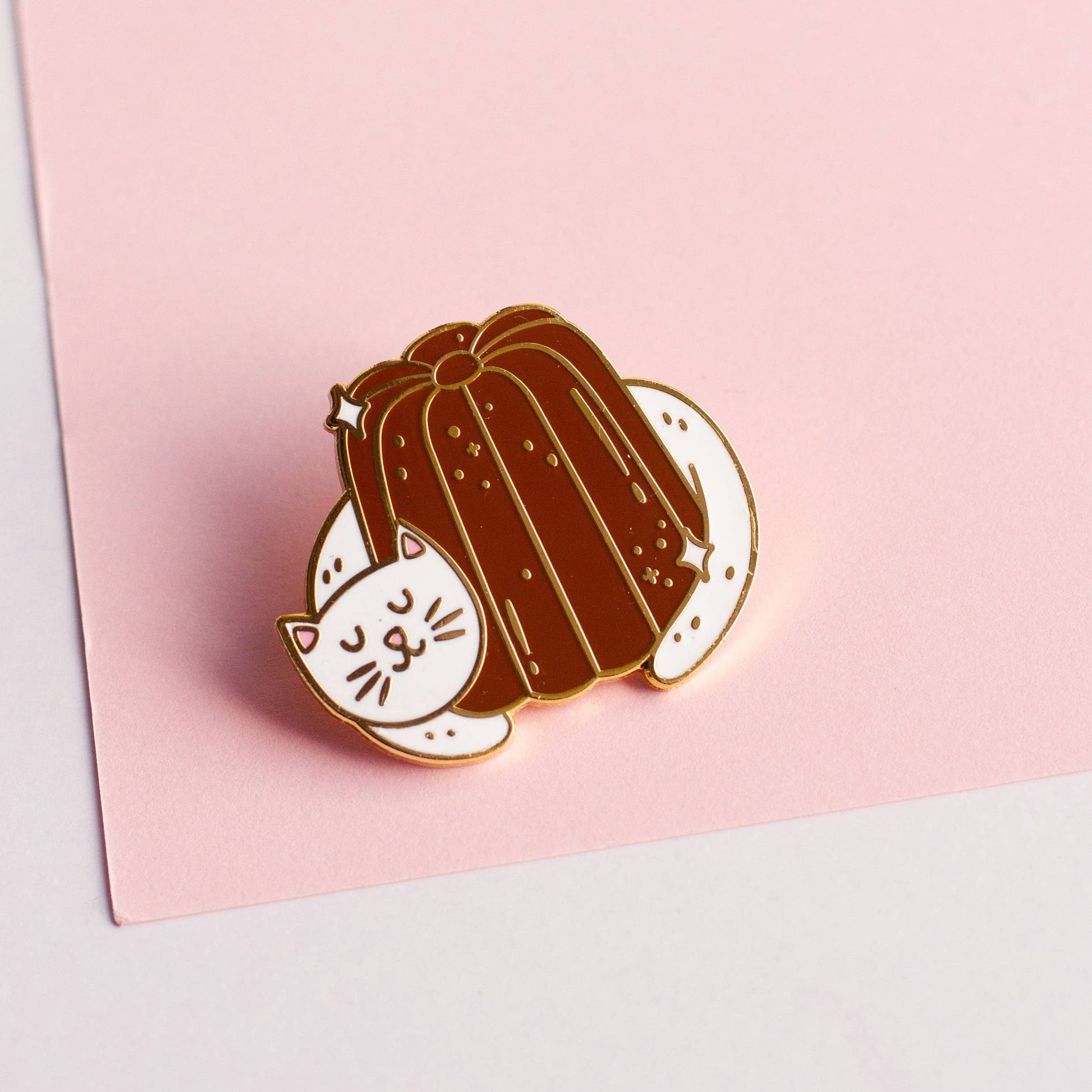 Pink paper background. Cat hugging a canele lapel pin.