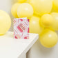 Greeting card with illustration of slices of pink birthday cake and candles on white background. Text "celebrating you is a piece of cake" is frosted onto the side of a piece of cake. Shown displayed on a white table with yellow balloons in the background.