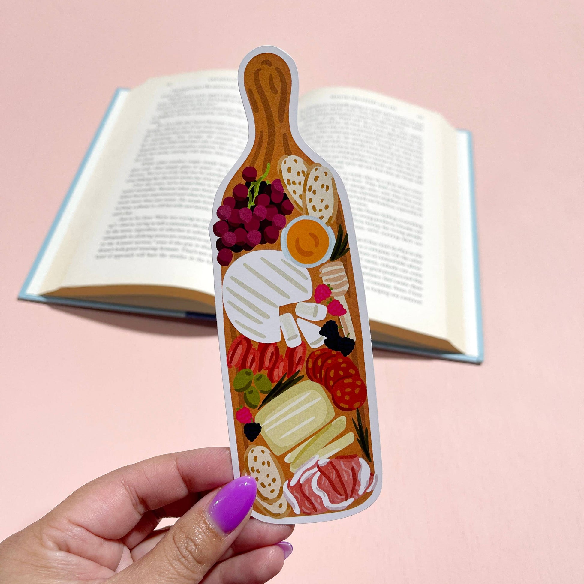 Hand holding a decorative bookmark shaped like a charcuterie board with food illustrations against an open book on a pink background