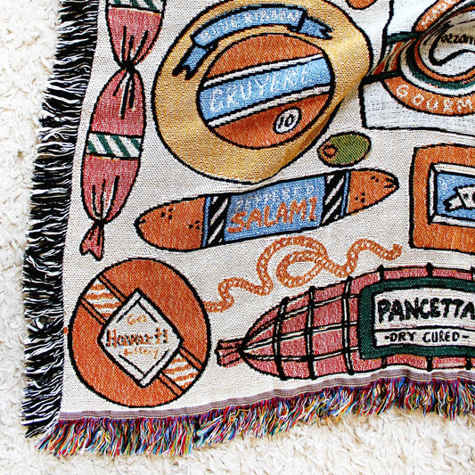 Cheese and Charcuterie tapestry blanket with assorted meats, cheeses, fruit, olives and deli wrapped items. Close up photo to show detail of fabric