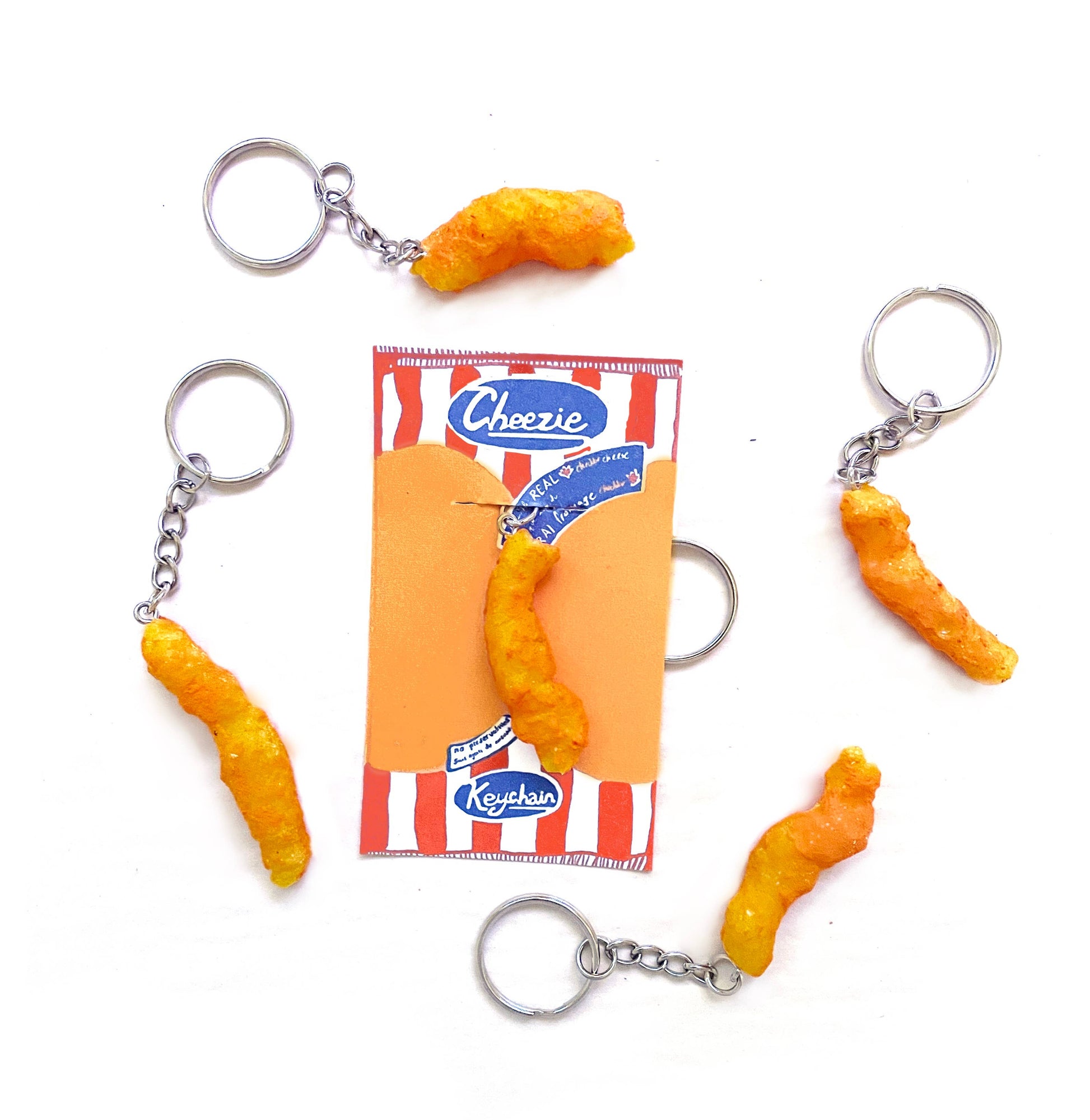 Keychain designed to look like a Canadian "Cheezie" snack hanging from a silver metal chain and keyring. Shown on red and white striped paper backing.
