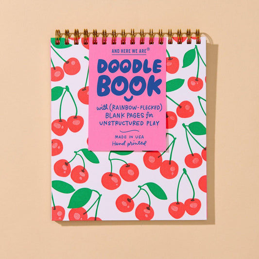 A spiral-bound Doodle Book with a white cover decorated with a red cherry pattern and a bright pink title block, photographed against a solid beige background.