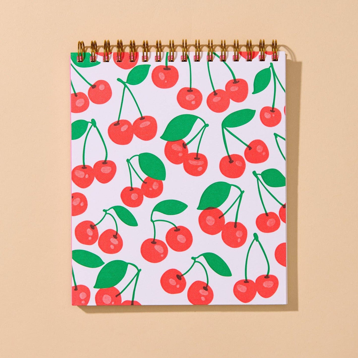 A spiral-bound notebook with a white cover decorated with a repeating pattern of red cherries and green leaves, displayed against a solid beige background.