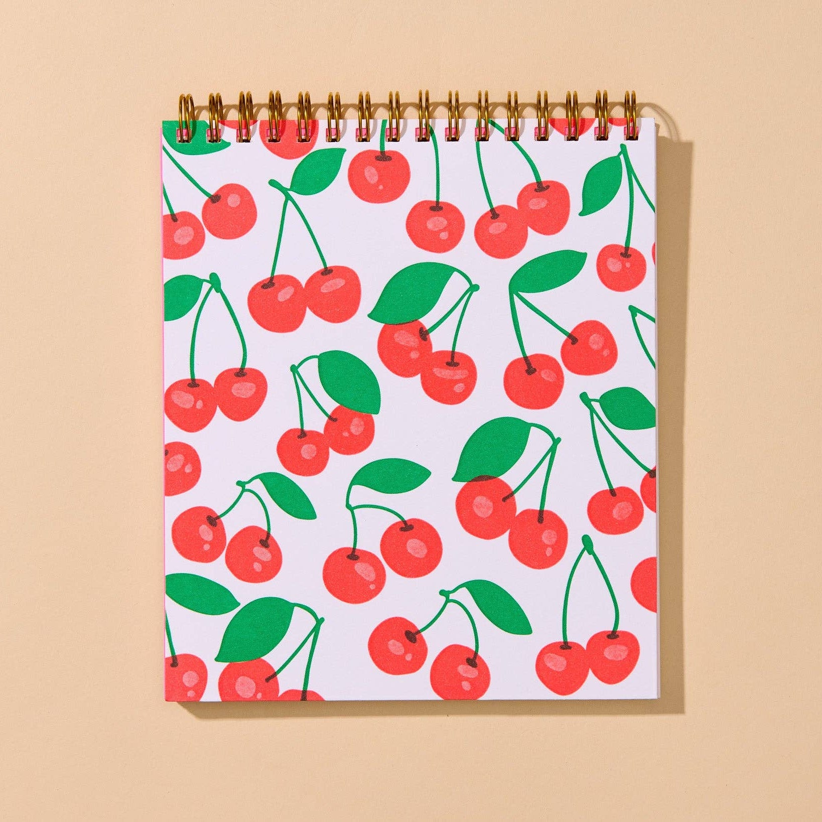A spiral-bound notebook with a white cover decorated with a repeating pattern of red cherries and green leaves, displayed against a solid beige background.