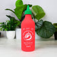 Lamp that looks like a chili sauce bottle, red bottle with green cap, on a white surface with plants in the background