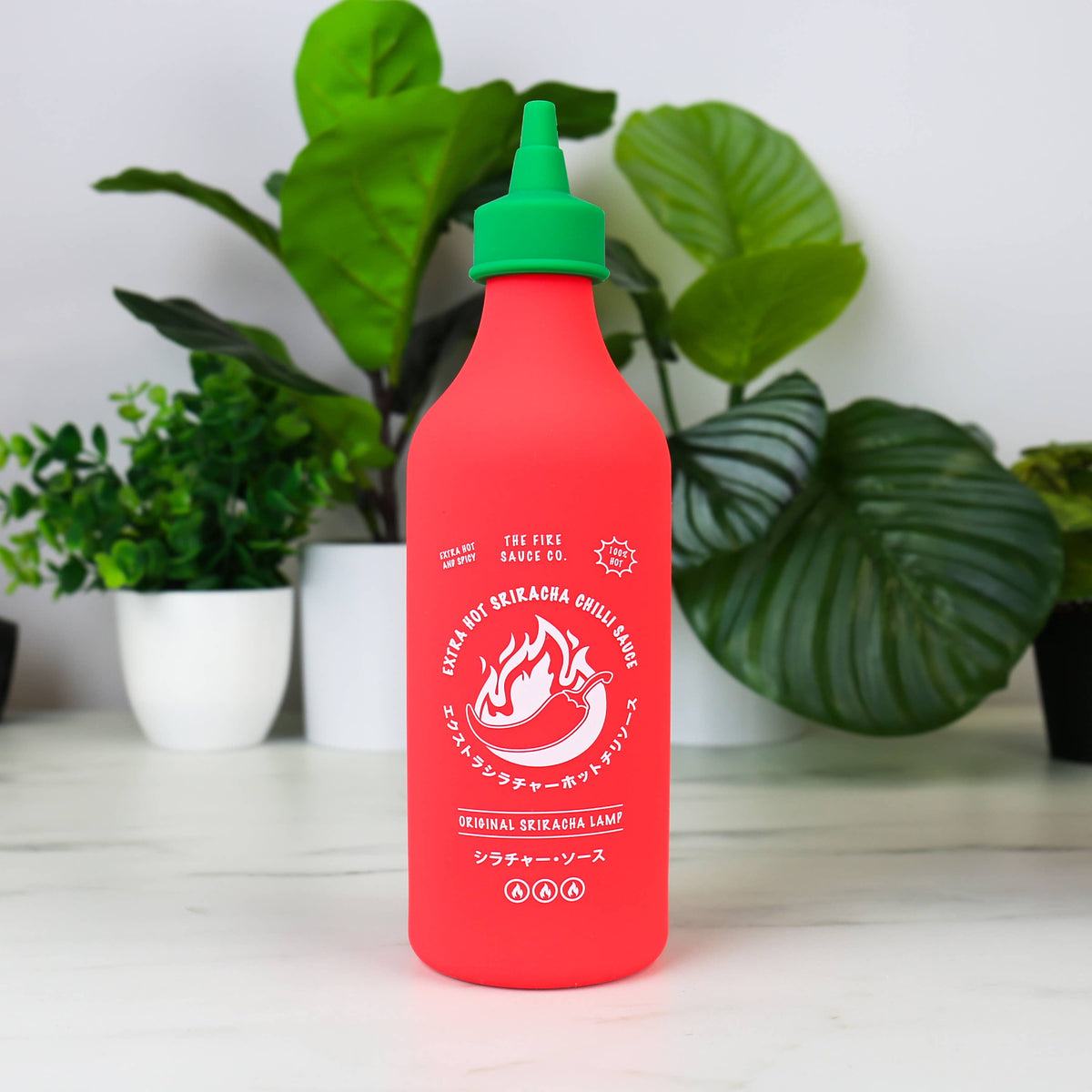 Lamp that looks like a chili sauce bottle, red bottle with green cap, on a white surface with plants in the background