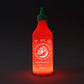 A novelty bottle lamp designed to look like a Sriracha sauce bottle, glowing with a warm red-orange light against a dark background.