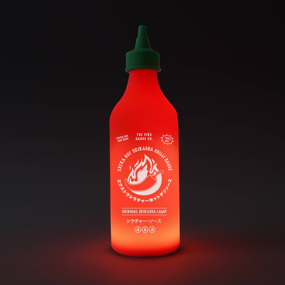 A novelty bottle lamp designed to look like a Sriracha sauce bottle, glowing with a warm red-orange light against a dark background.