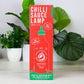 A tall red and green box of Chilli Sauce Lamp hot sauce stands upright on a white surface, with potted green plants in the background.