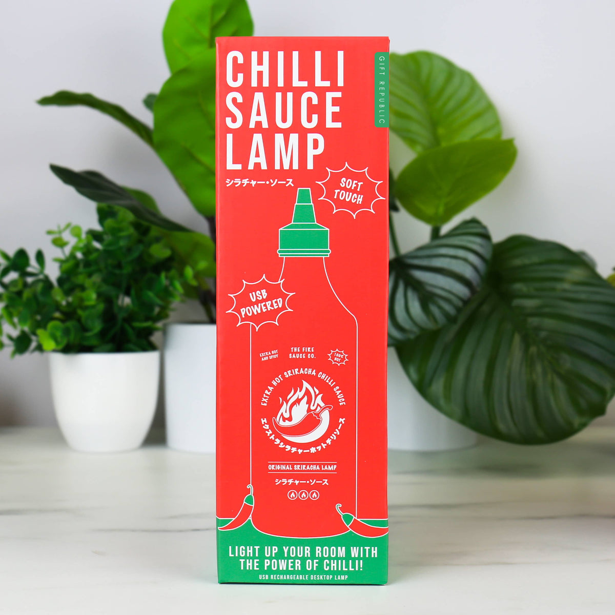 A tall red and green box of Chilli Sauce Lamp hot sauce stands upright on a white surface, with potted green plants in the background.