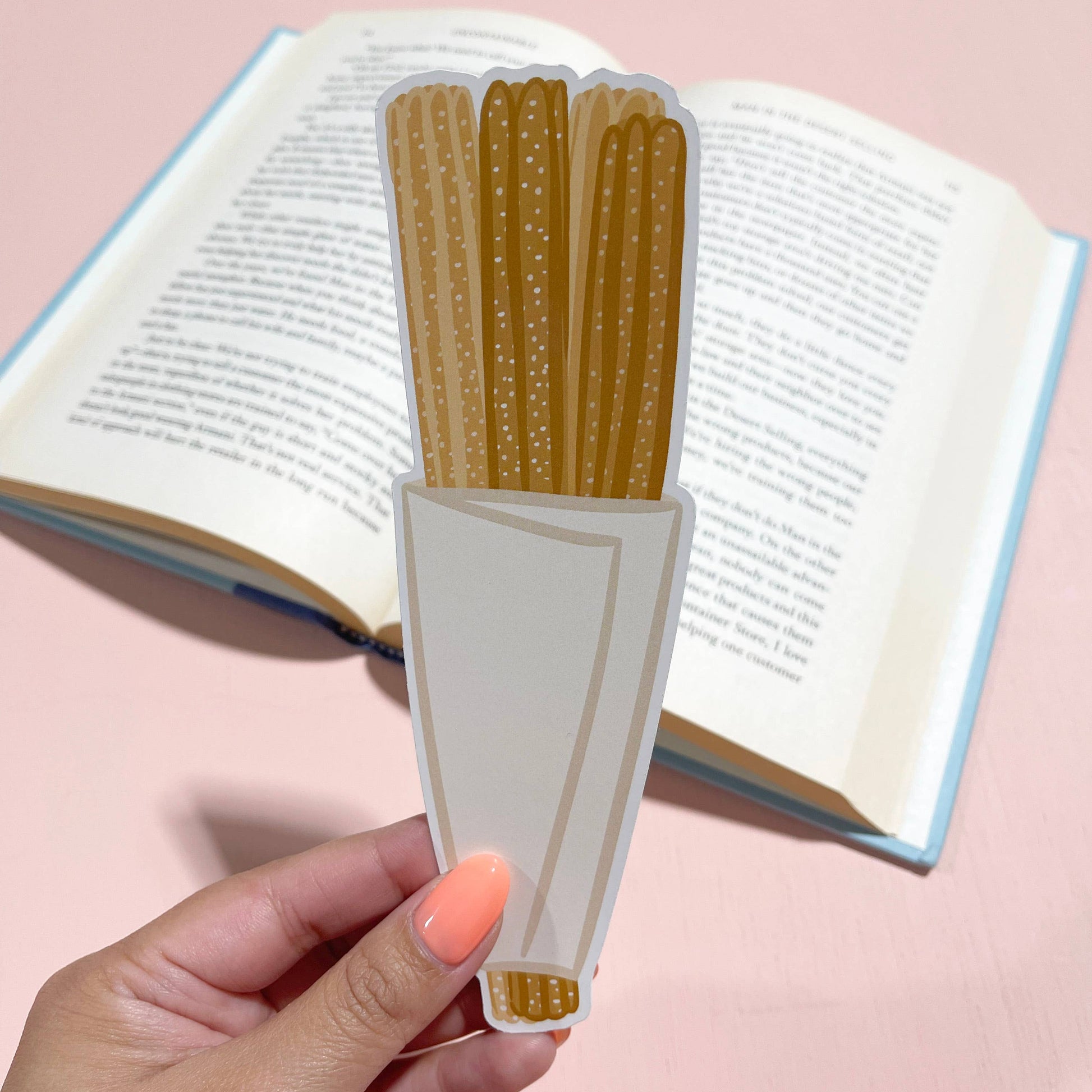 Hand holding a book with a unique bookmark shaped like churros against a pink background