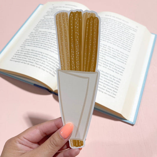 Hand holding a book with a unique bookmark shaped like churros against a pink background