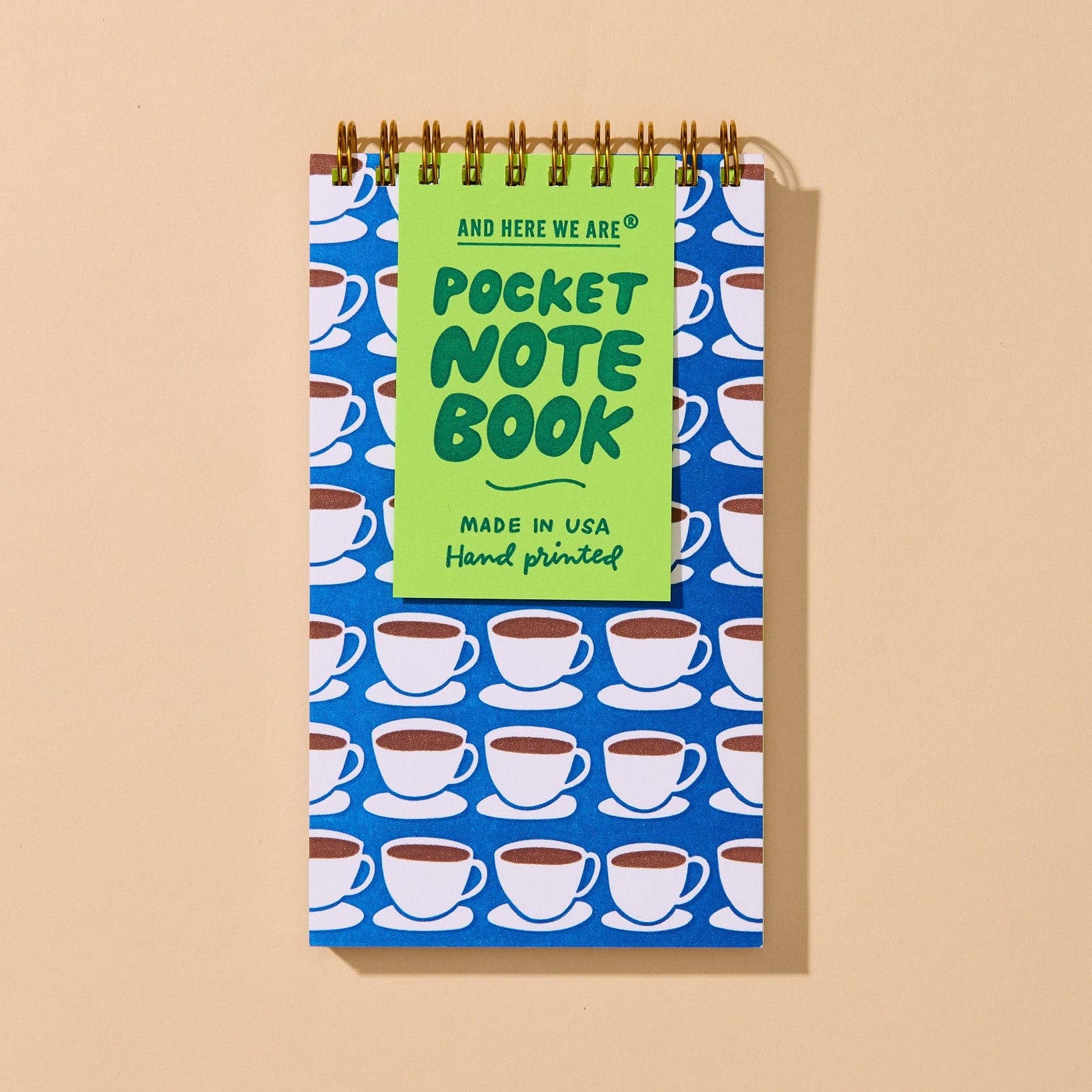 A spiral-bound pocket notebook with a blue cover patterned with white coffee cups and a bright green title panel reading 'Pocket Note Book' and 'And Here We Are Made in USA Hand printed,' shown on a warm beige background.
