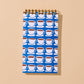 A spiral-bound notebook with a blue cover featuring a repeating pattern of white coffee cups on saucers, displayed against a solid beige background.