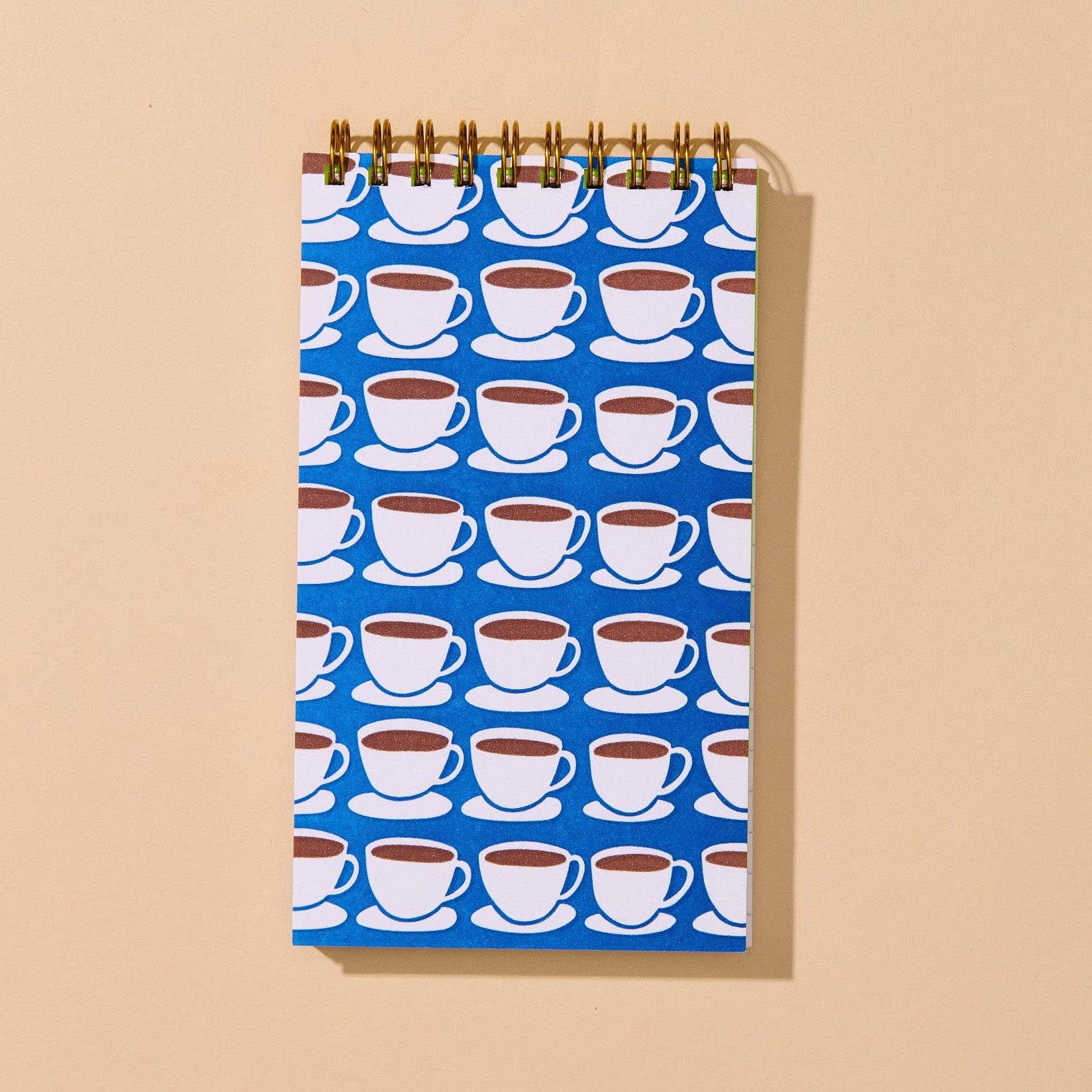 A spiral-bound notebook with a blue cover featuring a repeating pattern of white coffee cups on saucers, displayed against a solid beige background.
