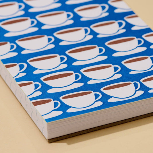A close-up of a notebook or journal with a blue cover patterned with repeating white coffee cups and saucers, resting on a light beige surface.