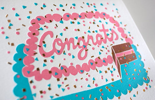 Congrats sheet cake card with sprinkles and confetti. The cake has a slice taken out of it to reveal chocolate cake with pink frosting. Close up photo to show the foil detail of the confetti.