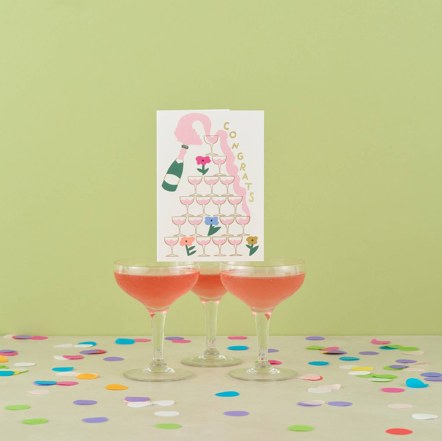 Three cocktail glasses with pink drinks on a table with confetti, next to a 'Congrats' card with champagne and flowers.
