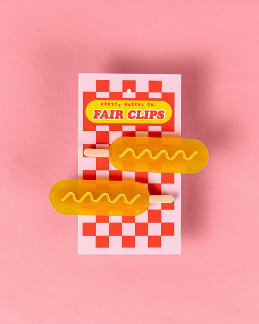 Two hairclips shaped like corndogs shown attached to red and white checkered backing board with text "Pretty Useful Co. - Fair Clips"