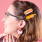 Woman in glasses wearing a corndog hair clip and a hotdog hair clip.