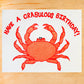 A square white greeting card or print featuring a red crab illustration and the text 'HAVE A CRABULOUS BIRTHDAY!' in red, placed on a light wood surface.