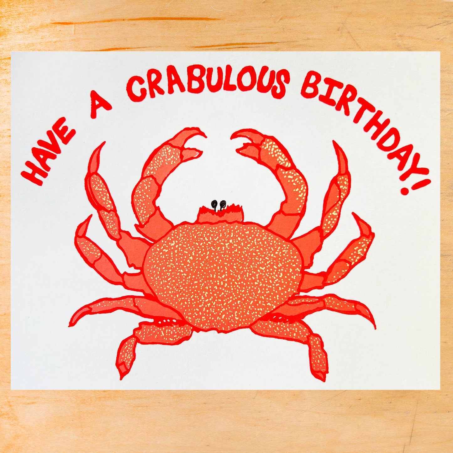 A square white greeting card or print featuring a red crab illustration and the text 'HAVE A CRABULOUS BIRTHDAY!' in red, placed on a light wood surface.