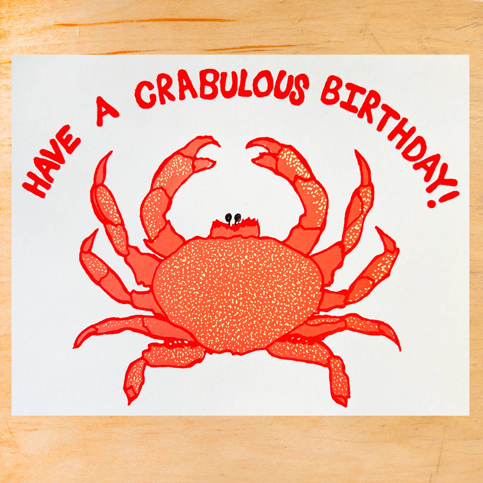 A square white greeting card or print featuring a red crab illustration and the text 'HAVE A CRABULOUS BIRTHDAY!' in red, placed on a light wood surface.