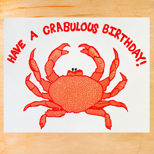 A square white greeting card or print featuring a red crab illustration and the text 'HAVE A CRABULOUS BIRTHDAY!' in red, placed on a light wood surface.